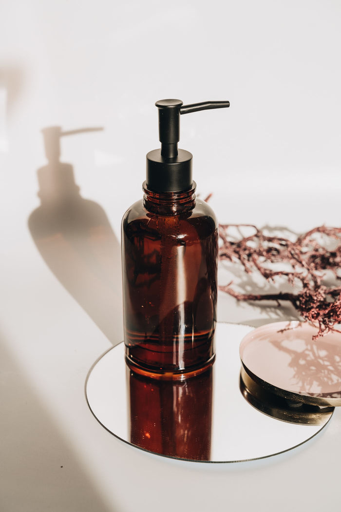 Amber glass pump soap dispenser on a round mirror in sunlight with shadow on white wall