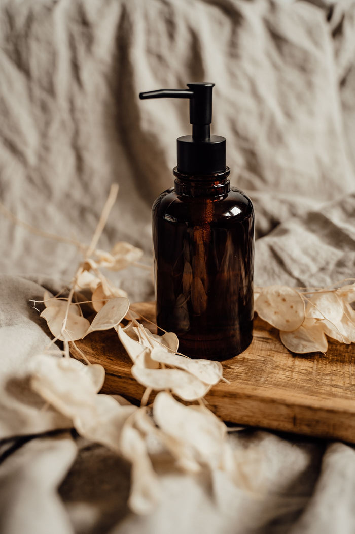 Amber glass soap dispenser on a wooden board with dried flowers and beige linen