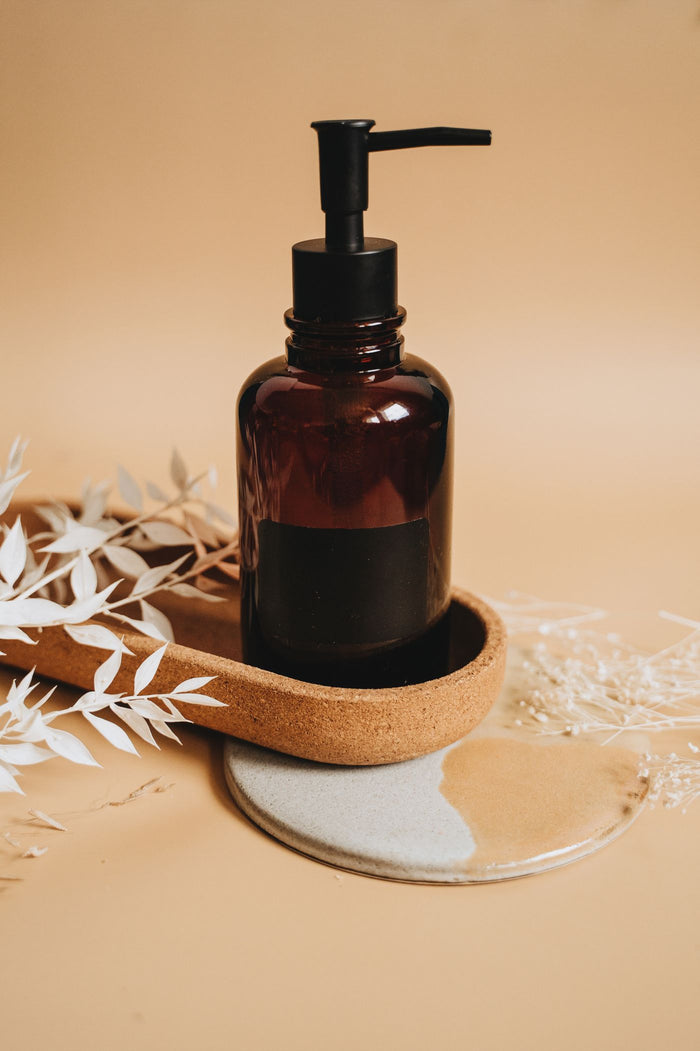 Amber glass pump soap dispenser with blank black band in a cork tray with dried foliage