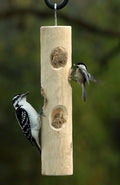 Woodpecker and chickadee eat at a cedar log feeder, with holes containing suet plugs. Feeder hangs from silver hook.
