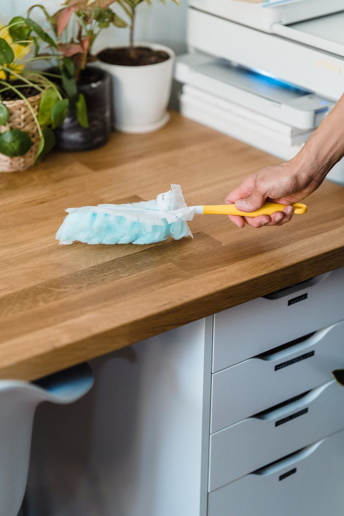 Pale blue microfiber duster with a yellow handle sweeping a light wooden desktop