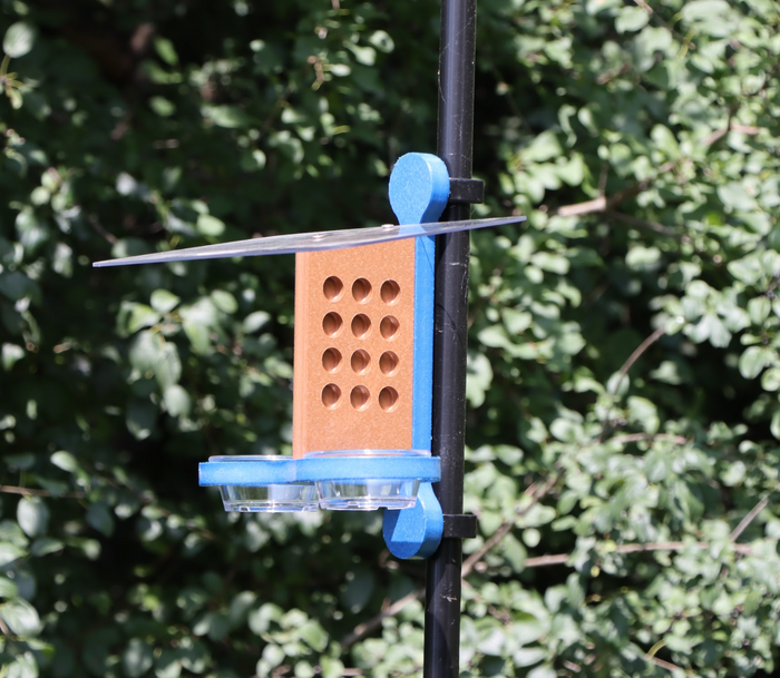 A bird feeder clipped to a black pole, featuring a brown center with circular holes, a blue mounting bracket and base, black plastic clips, a clear roof and two small clear bowls, set against a background of green foliage.