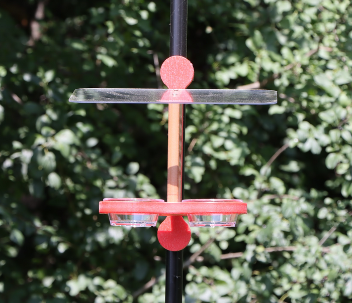 Straight on view of a bird feeder clipped to a black pole featuring a brown center, a red mounting bracket and base, black plastic clips, a clear roof, and two small clear bowls, set against a background of green foliage.