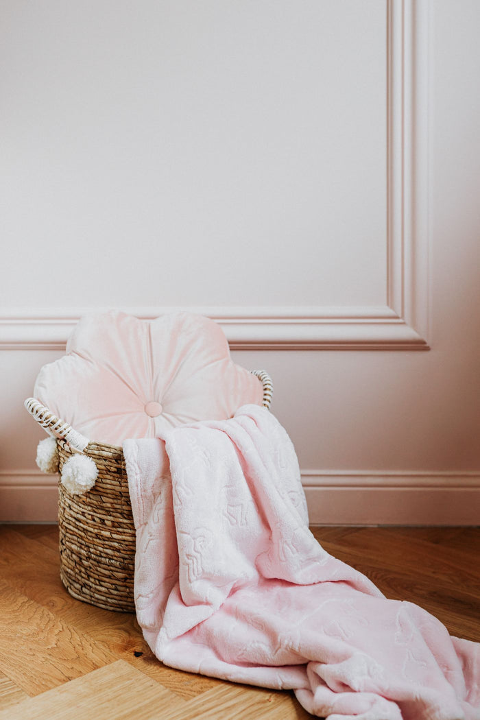 Natural woven storage basket with white pom-pom trim on a wooden floor, filled with a pink throw blanket and round cushion